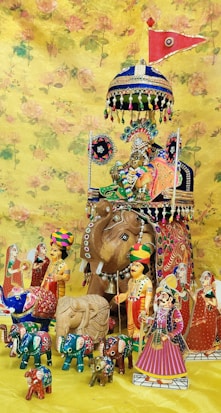 A colorful arrangement of traditional Indian dolls and figurines is displayed against a floral yellow backdrop. The centerpiece features a large wooden elephant adorned with intricate designs, topped with a vibrant umbrella and a red flag. Surrounding the elephant are various figures, including human characters dressed in traditional attire, and smaller elephants painted in bright colors.