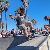 A skateboarder performs a trick on the edge of a skatepark ramp, surrounded by palm trees under a clear blue sky. In the background, there are other people, some of whom appear to be watching and others engaged in their activities.