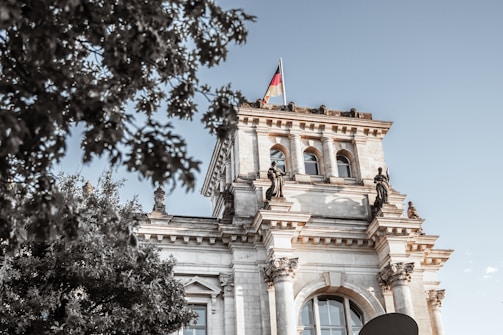 A classical building with ornate architecture featuring columns and statues is seen, with a German flag flying at the top. The building appears to be part of a historic or governmental structure. In the foreground, there are tree branches with green leaves.