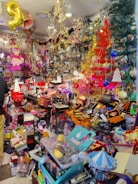 A colorful and crowded store filled with various items including toys, decorations, and gift boxes. The shelves are packed with different objects, and vibrant holiday decorations such as tinsel and Christmas trees are visible. A pink doll hangs from the ceiling, and numerous brightly colored boxes and products are scattered throughout the space.
