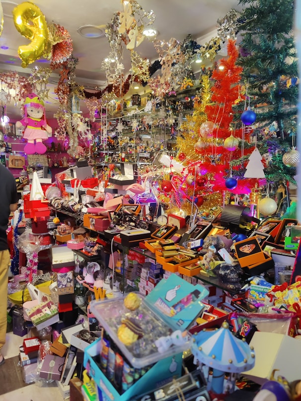 A colorful and crowded store filled with various items including toys, decorations, and gift boxes. The shelves are packed with different objects, and vibrant holiday decorations such as tinsel and Christmas trees are visible. A pink doll hangs from the ceiling, and numerous brightly colored boxes and products are scattered throughout the space.