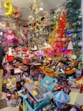 A colorful and crowded store filled with various items including toys, decorations, and gift boxes. The shelves are packed with different objects, and vibrant holiday decorations such as tinsel and Christmas trees are visible. A pink doll hangs from the ceiling, and numerous brightly colored boxes and products are scattered throughout the space.