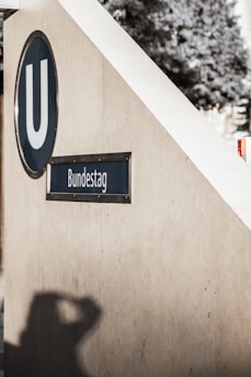 An entrance to a subway station with a sign reading 'Bundestag'. The wall has a distinct shadow of a person captured in the photograph. There is a 'U' symbol indicating a subway entrance. The background appears to have trees or foliage, suggesting an outdoor setting.