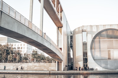 A modern architectural scene featuring a wide bridge connecting two contemporary buildings. On the left, a cyclist and a pedestrian are seen along a riverbank lined with trees and a stone wall. The right side displays a group of people gathered near a distinctive building with large circular windows, reflecting a minimalist design.