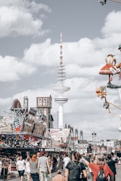 A bustling amusement park scene with numerous people walking, engaging with various attractions and rides. A prominent radio tower stands tall in the background, surrounded by rides and festive decorations. The scene captures a lively atmosphere under a partly cloudy sky.