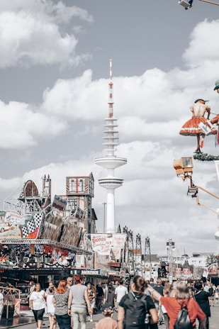 A bustling amusement park scene with numerous people walking, engaging with various attractions and rides. A prominent radio tower stands tall in the background, surrounded by rides and festive decorations. The scene captures a lively atmosphere under a partly cloudy sky.