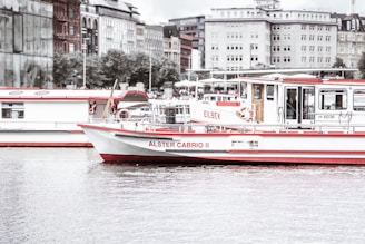 A river boat named Alster Cabrio II is floating on calm waters. The boat has a white hull with contrasting red accents. In the background, there are multiple buildings with different architectural styles, and another boat is visible parked adjacent to the featured one. The sky appears overcast, creating a calm and muted atmosphere.