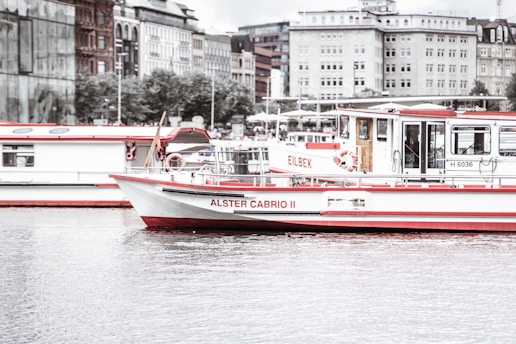 A river boat named Alster Cabrio II is floating on calm waters. The boat has a white hull with contrasting red accents. In the background, there are multiple buildings with different architectural styles, and another boat is visible parked adjacent to the featured one. The sky appears overcast, creating a calm and muted atmosphere.