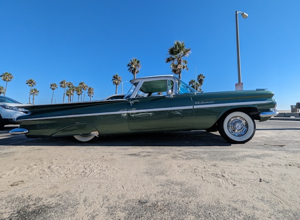 A classic green car is parked near a beach with palm trees in the background. The car has a sleek design, featuring white-wall tires and shiny chrome accents. The sky is clear and bright blue, and the area appears sunny and open, adding to the vintage and laid-back atmosphere.