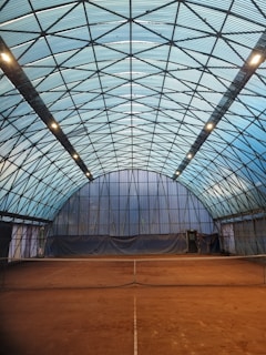 An indoor tennis court with a high, arched ceiling made of translucent panels supported by a metal framework. The court surface appears to be clay, characterized by its reddish-brown color. Lighting is provided by a series of row lights attached to the ceiling trusses, casting a soft glow over the area. A net divides the court in the middle, and the back wall has a large fabric or tarp hanging, possibly to prevent balls from leaving the court.
