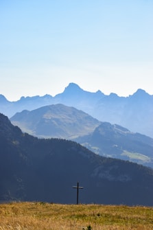 A serene landscape featuring a cross in the foreground set against a backdrop of rolling, forested hills and majestic mountains. The sky is clear and bright, amplifying the peacefulness of the scene.