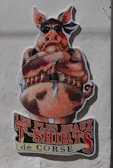 A large, colorful cartoon illustration of a pig, wearing a black bandana and sunglasses, positioned against a light-colored wall. The pig is depicted with a stern expression, arms crossed, and an ammunition belt across its chest. Beneath the illustration, bold text in French reads 'Les Plus Beaux T-Shirts de Corse,' referencing a clothing theme.