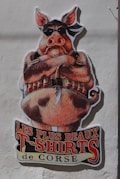 A large, colorful cartoon illustration of a pig, wearing a black bandana and sunglasses, positioned against a light-colored wall. The pig is depicted with a stern expression, arms crossed, and an ammunition belt across its chest. Beneath the illustration, bold text in French reads 'Les Plus Beaux T-Shirts de Corse,' referencing a clothing theme.