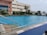 An outdoor swimming pool with clear blue water is surrounded by greenery and palm trees. Adjacent is a multi-story building with large windows. The pool's edge features metal handrails and warning signs on the floor, indicating caution for slippery surfaces. The sky is mostly clear, suggesting a pleasant day.