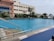 An outdoor swimming pool with clear blue water is surrounded by greenery and palm trees. Adjacent is a multi-story building with large windows. The pool's edge features metal handrails and warning signs on the floor, indicating caution for slippery surfaces. The sky is mostly clear, suggesting a pleasant day.