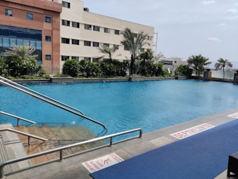 An outdoor swimming pool with clear blue water is surrounded by greenery and palm trees. Adjacent is a multi-story building with large windows. The pool's edge features metal handrails and warning signs on the floor, indicating caution for slippery surfaces. The sky is mostly clear, suggesting a pleasant day.