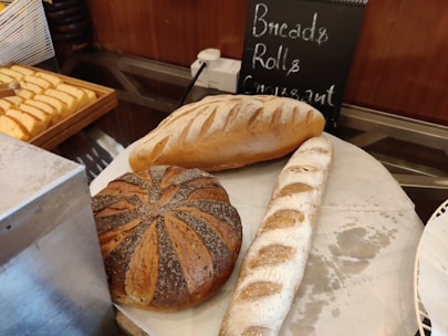 A display of various types of bread placed on a counter, including a loaf of white bread, a round whole grain loaf, and a baguette. There is a chalkboard sign in the background listing 'Bread, Rolls, Croissant' and a tray of sliced bread on the side.