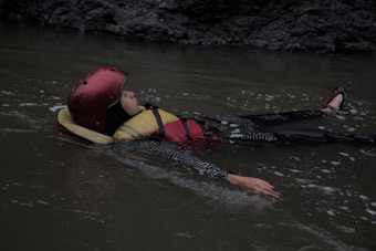 A person floats on calm water, wearing a red helmet and life jacket. The surrounding environment is dimly lit, suggesting an outdoor location with rocky edges.