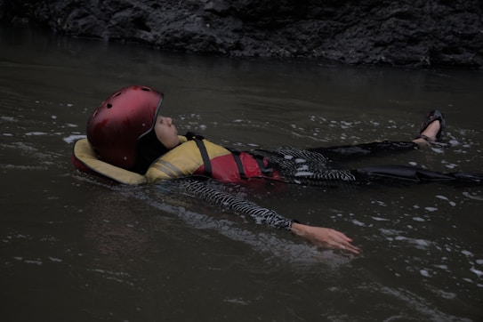 A person floats on calm water, wearing a red helmet and life jacket. The surrounding environment is dimly lit, suggesting an outdoor location with rocky edges.