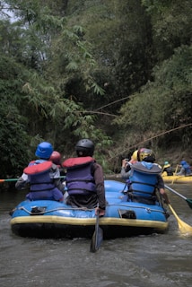 A group of people wearing helmets and life jackets are seated in a blue inflatable raft on a river. The surrounding environment is lush with dense green foliage, suggesting a forested area. Some individuals are holding paddles, indicating they are engaging in rafting.