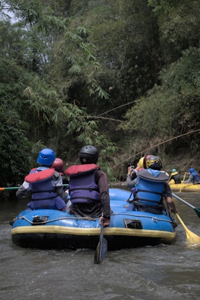 A group of people wearing helmets and life jackets are seated in a blue inflatable raft on a river. The surrounding environment is lush with dense green foliage, suggesting a forested area. Some individuals are holding paddles, indicating they are engaging in rafting.