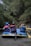 A group of people wearing helmets and life jackets are seated in a blue inflatable raft on a river. The surrounding environment is lush with dense green foliage, suggesting a forested area. Some individuals are holding paddles, indicating they are engaging in rafting.