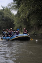 A group of people wearing life jackets and helmets are paddling an inflatable raft down a river. The river is surrounded by lush green foliage, with overhanging trees on both sides. Each person holds a paddle, and they are all focused on navigating through the water.
