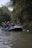 A group of people wearing life jackets and helmets are paddling an inflatable raft down a river. The river is surrounded by lush green foliage, with overhanging trees on both sides. Each person holds a paddle, and they are all focused on navigating through the water.