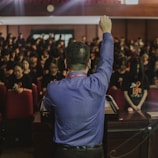 A person stands at a podium facing a large audience, raising their fist in a gesture of encouragement or solidarity. The audience is seated, consisting mainly of people wearing dark shirts with some form of insignia. The setting appears to be a formal or professional gathering, possibly in an auditorium or conference room.