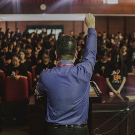 A person stands at a podium facing a large audience, raising their fist in a gesture of encouragement or solidarity. The audience is seated, consisting mainly of people wearing dark shirts with some form of insignia. The setting appears to be a formal or professional gathering, possibly in an auditorium or conference room.