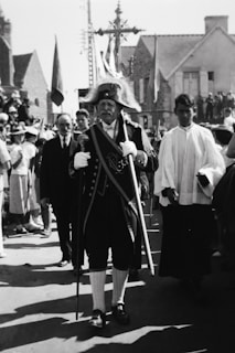 A historical scene with a man dressed in ceremonial attire and a feathered hat procession through a street. He carries a staff and wears a coat adorned with embellishments. Several people surround him, some in ceremonial robes, and there are onlookers in the background. The architecture suggests an old European setting.