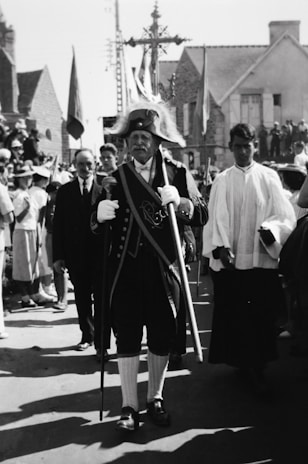 A historical scene with a man dressed in ceremonial attire and a feathered hat procession through a street. He carries a staff and wears a coat adorned with embellishments. Several people surround him, some in ceremonial robes, and there are onlookers in the background. The architecture suggests an old European setting.