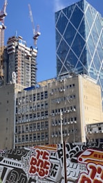 Several tall buildings under construction and completed high-rise structures are present. The scene includes cranes and scaffolding with a modern glass building featuring a crisscross pattern. A concrete building in the foreground is covered in numerous air conditioning units. In the lower part of the image, there is a colorful graffiti-covered barrier with stylized text.