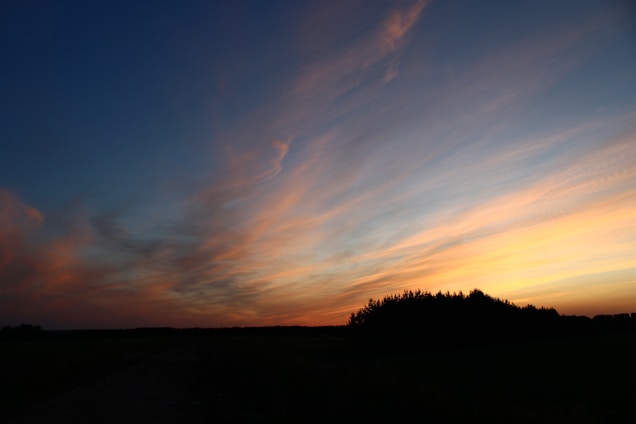 The image captures a serene landscape during sunset. The sky is painted with vibrant hues of orange, pink, and blue, creating a captivating gradient. Wispy clouds stretch across the sky, adding texture to the scene. Below, the silhouette of a tree line is visible, framing the lower edge of the image.