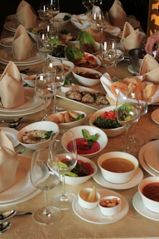 A lavish banquet table set elegantly with folded napkins and multiple glassware, surrounded by a variety of dishes including soups, salads, dips, breads, and other appetizers. The arrangement is inviting and suggests a formal dining occasion.