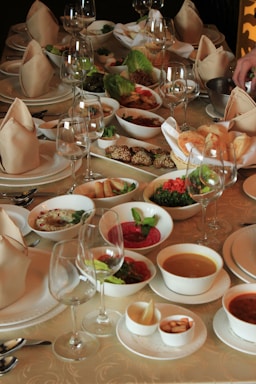 A lavish banquet table set elegantly with folded napkins and multiple glassware, surrounded by a variety of dishes including soups, salads, dips, breads, and other appetizers. The arrangement is inviting and suggests a formal dining occasion.