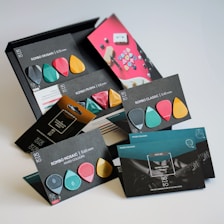 A collection of colorful guitar picks arranged with their packaging, displaying different shapes and thickness options. The packaging is stylish with a modern design, featuring various colors like teal, pink, gold, and black. Text describing each pick style such as Rombo Origami, Rombo Prisma, and Rombo Classic is visible.