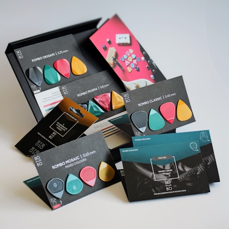 A collection of colorful guitar picks arranged with their packaging, displaying different shapes and thickness options. The packaging is stylish with a modern design, featuring various colors like teal, pink, gold, and black. Text describing each pick style such as Rombo Origami, Rombo Prisma, and Rombo Classic is visible.