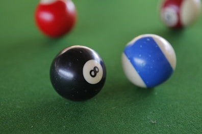 A close-up view of billiard balls on a green pool table surface, prominently featuring a black number 8 ball and a blue and white striped ball in focus, with blurred red and white balls in the background.