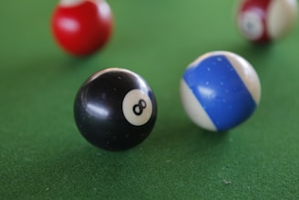 A close-up view of billiard balls on a green pool table surface, prominently featuring a black number 8 ball and a blue and white striped ball in focus, with blurred red and white balls in the background.