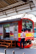 A train is stationary at a platform with a worker standing beside it. The train has a red and silver exterior with yellow and red stripes. There are two small flags displayed near the front of the train. Overhead, structural metal beams and parts of the station roof are visible.