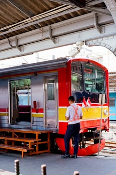 A train is stationary at a platform with a worker standing beside it. The train has a red and silver exterior with yellow and red stripes. There are two small flags displayed near the front of the train. Overhead, structural metal beams and parts of the station roof are visible.