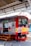 A train is stationary at a platform with a worker standing beside it. The train has a red and silver exterior with yellow and red stripes. There are two small flags displayed near the front of the train. Overhead, structural metal beams and parts of the station roof are visible.