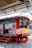 A train is stationary at a platform with a worker standing beside it. The train has a red and silver exterior with yellow and red stripes. There are two small flags displayed near the front of the train. Overhead, structural metal beams and parts of the station roof are visible.
