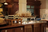 A bakery display in a rustic-style interior featuring shelves with packaged bread and jars. There are stacks of yogurt containers on wooden risers, and an assortment of items in a glass display counter. The surrounding area has warm wooden cabinetry.