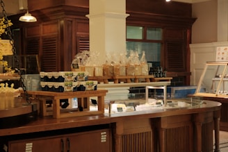 A bakery display in a rustic-style interior featuring shelves with packaged bread and jars. There are stacks of yogurt containers on wooden risers, and an assortment of items in a glass display counter. The surrounding area has warm wooden cabinetry.