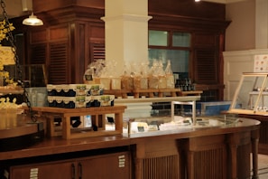 A bakery display in a rustic-style interior featuring shelves with packaged bread and jars. There are stacks of yogurt containers on wooden risers, and an assortment of items in a glass display counter. The surrounding area has warm wooden cabinetry.