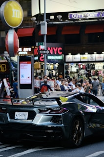 A sleek, modern sports car with a convertible roof and a New York license plate is parked on a busy city street. Surrounding the car are numerous pedestrians, a mix of locals and tourists. The background includes various storefronts and neon signs, with an 'NYC Mega Mart' and an 'm&m' logo prominently visible. The scene is vibrant with lots of activity and a bustling urban atmosphere.