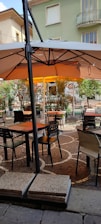 An outdoor café setting features tables and chairs arranged on a decorative cobblestone patio. There is a large beige sun umbrella providing shade over the seating area. Potted plants and greenery border the patio, and in the background, part of a residential building with balconies is visible.