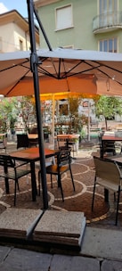 An outdoor café setting features tables and chairs arranged on a decorative cobblestone patio. There is a large beige sun umbrella providing shade over the seating area. Potted plants and greenery border the patio, and in the background, part of a residential building with balconies is visible.